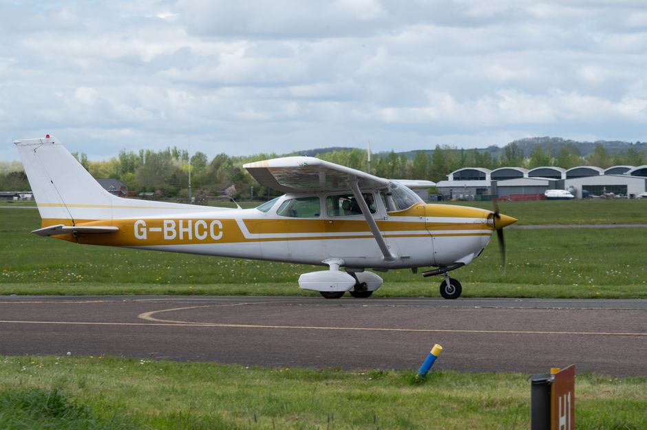 A small, white aircraft with yellow and gray markings, bearing the registration G-BHCC, positioned on a runway at Biggin Hill Airport. The aircraft features a high-wing design with a single propeller 