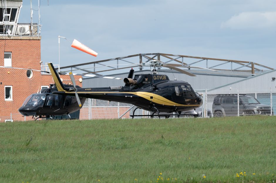 A black and gold helicopter parked on a grassy area adjacent to a modern building with a brick section and large metallic hangar structure. The helicopter has a sleek, polished appearance with visible