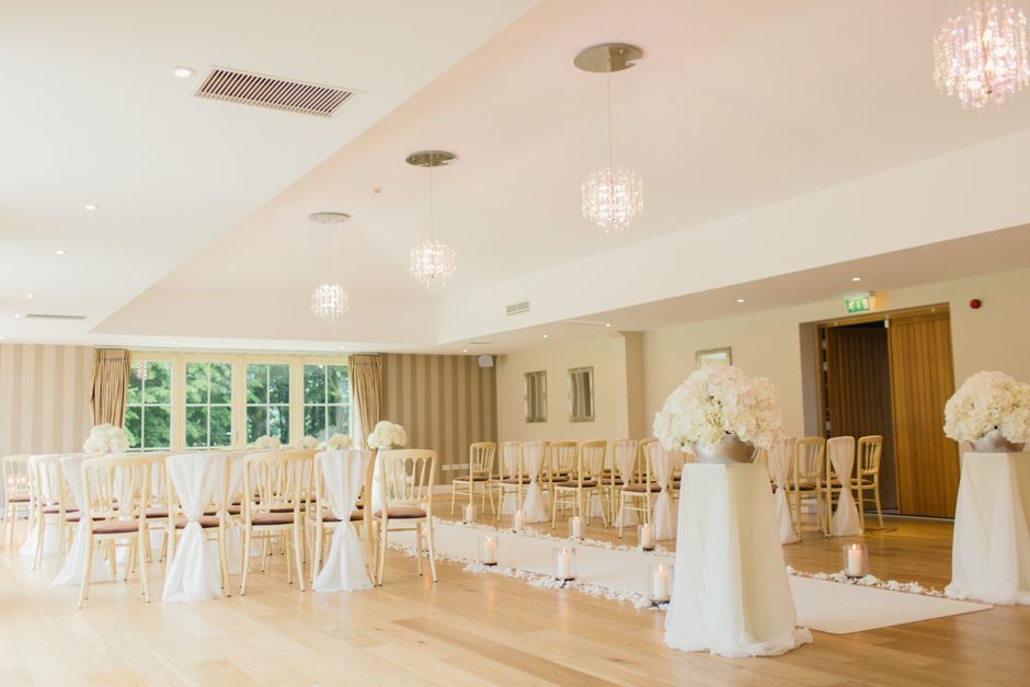 An elegant wedding ceremony setup inside a bright, softly lit room featuring large windows with greenery views. The arrangement includes cream-colored chairs with white sashes, positioned around a whi