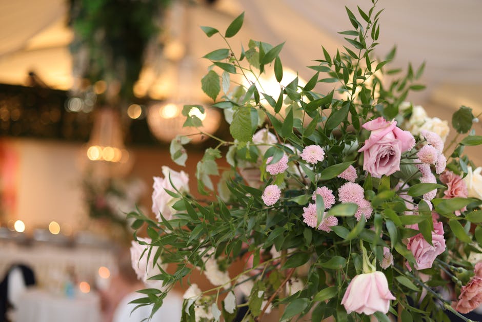 A close-up of a floral arrangement featuring soft pink roses and small pink pompon chrysanthemums, complemented by lush green leaves and sprigs, arranged in a natural, loose style. The bouquet is set 