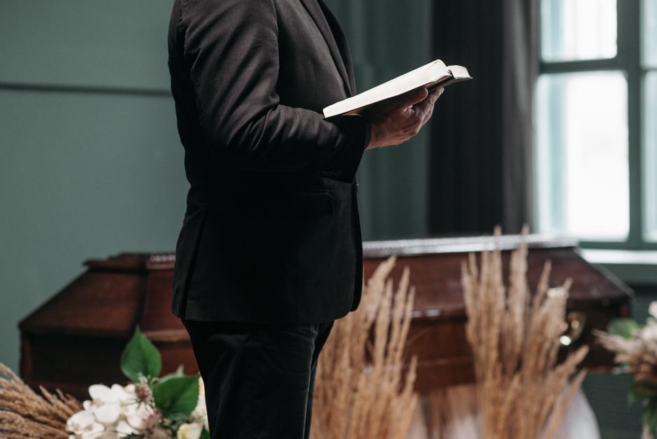 A person dressed in a black suit holding an open book, standing indoors with a soft natural light coming through a window. Behind them, there is a wooden casket surrounded by floral arrangements featu
