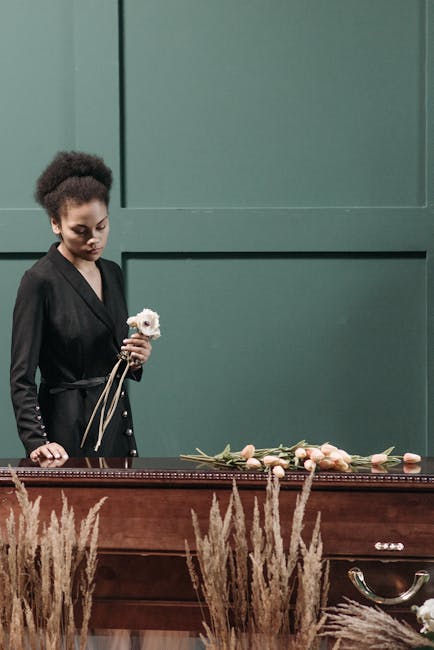 A woman with dark, curly hair styled in an updo, dressed in a black blazer, stands beside a wooden table holding a small white flower. The table is decorated with a floral arrangement featuring pale p