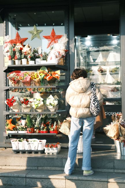 A woman dressed in a beige puffer jacket and light blue jeans stands on the pavement in front of a flower shop on Biggin Hill High Street, observing a display of floral arrangements and gifts through 