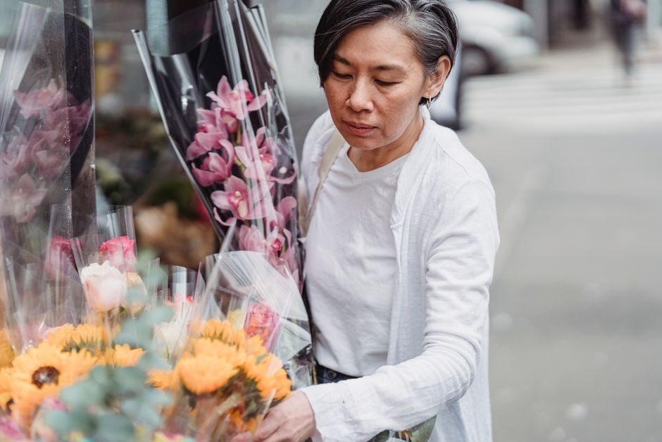 A woman with short gray hair, dressed in a white top and light jacket, examines a vibrant display of fresh flower bouquets outside a floral shop. She is holding several large bouquets wrapped in clear