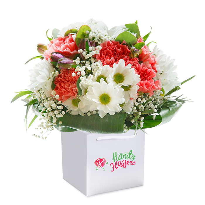 Presented against a flawless white background, this floral composition radiates freshness-an image drawn from the heart of Biggin Hill, capturing the spirit of the community. Centered in a square white gift bag with practical ribbon handles, the bouquet itself is a layered symphony of texture and colour: lush coral and soft peach carnations unfurl with gentle dynamism, their numerous ruffled petals inviting touch and admiration. Clustered beside them are crisp white daisy-like blossoms, each sporting a vivid green center, injecting cheer as bright and clear as an open day at Biggin Hill Recreation Ground. Ethereal plumes of baby's breath float in and out of view, filling the spaces between and contributing an airy, almost dreamy softness. Tucked discreetly along the left rim, purple-edged buds-perhaps alstroemeria not yet fully open-hint at the unfolding of new chapters or occasions soon to be celebrated. Deep, verdant leaves provide a glossy, natural collar, grounding the arrangement with lushness and stability. The white bag-which could as easily be found in the hands of someone leaving a local Biggin Hill bakery-bears no distraction, keeping the attention on florals and craftsmanship. The entire scene is illuminated with gentle, even light, allowing every colour to shine true. Perfectly suited for celebrations, warm wishes, or welcoming guests home from a walk to Saltbox Hill, this bouquet embodies thoughtful modern luxury in a distinctly Biggin Hill setting.