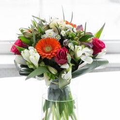This exceptional bouquet, styled by a local Biggin Hill florist, is an ode to the art of balancing vibrancy, form, and heartfelt emotion. The creamy peach and lively orange gerberas, their petals unfurling like the first warm rays over the Kentish hills, create a welcoming halo at the bouquet's core. Hidden among their gentle brightness are magenta roses-each bud a velvet spiral, deepening the arrangement's character and evoking the romance of cozy country evenings. Clusters of white alstroemeria, delicate spray roses, and whisper-soft pink blossoms offer a gentle lift, like morning clouds drifting past Leaves Green. The scene is anchored by robust, paddle-shaped aspidistra leaves, artfully layered and curled protectively around the base, forging a harmonious link between cultivated and wild beauty. Slender greenery threads throughout, adding a sense of abundance and vitality. All of this rises from a pristine glass vase, its transparency a symbol of freshness and authenticity. Light drapes softly over the composition, inviting the eye to linger on every detail while instilling a quiet cheer perfect for housewarmings or new journeys in Biggin Hill. There's an almost tangible sensation of crisp fragrance, blending floral sweetness and fresh green undercurrents. This floral ensemble is more than a bouquet-it's an embodiment of local care, ready to mark achievements, milestones, or simple everyday joys.